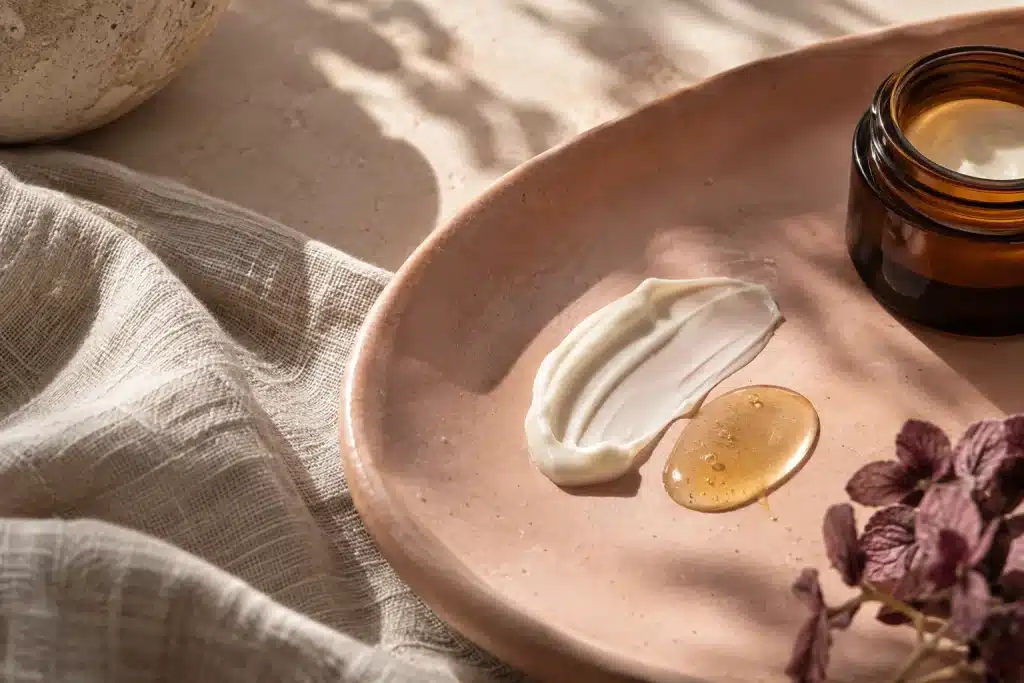 Bodegón editorial de crema marfil, sérum dorado y tarro ámbar sin etiqueta sobre cerámica rosa arcilla.