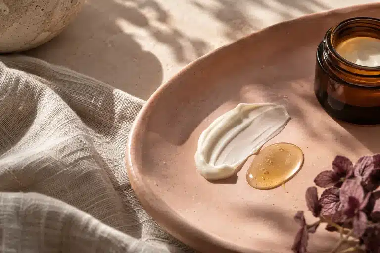 Bodegón editorial de crema marfil, sérum dorado y tarro ámbar sin etiqueta sobre cerámica rosa arcilla.